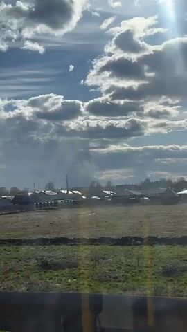The camera, likely from a moving vehicle, looks out over a rural landscape with scattered houses under a sky filled with cumulus clouds. The sun is bright, creating highlights and shadows across the scene. In the distance, what appears to be either very heavy rain or a column of smoke rises, partially obscuring the view of the houses. The foreground consists of fields with sparse vegetation. There are cell towers visible near the houses, suggesting a modern infrastructure within this rural setting. The overall impression is one of a dynamic weather event unfolding in a semi-urbanized rural area.
