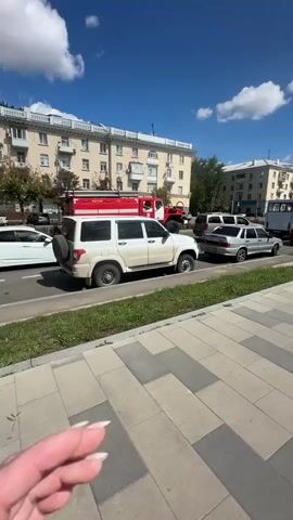 The video shows a street scene under a bright blue sky with white clouds. In the foreground, a sidewalk is paved with large gray and light gray rectangular tiles, bordered by a strip of green grass. To the left of the sidewalk, a road is visible with several cars. Closest to the camera is a white SUV parked on the side of the road, facing away from the camera, with a spare tire mounted on its rear. Further down the road, a silver sedan is also parked. Beyond these vehicles, a red fire truck with flashing lights is stopped, partially obscured by other vehicles. In the background, there are multi-story buildings with cream-colored facades. The overall impression is a street in an urban environment, possibly experiencing some traffic disruption due to the presence of the fire truck.