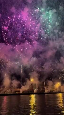 The video shows a fireworks display over a body of water at night. The fireworks are various colors, including pink, purple, and green, and are exploding in the dark sky. There is a low horizon line visible, likely a shoreline with some lights and possibly buildings. The water reflects the colors of the fireworks. The display appears to be a vibrant and active one with multiple explosions occurring simultaneously.