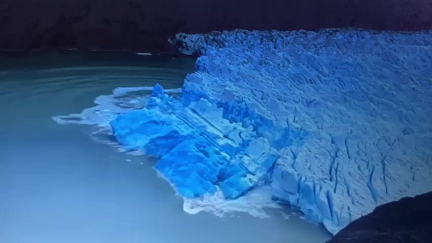 A glacial formation is visible in the video. The glacier is a large mass of ice, with sections of brilliant blue ice protruding into a body of water. The water appears somewhat murky and turbulent, with white foam or ice fragments around the edges of the glacier. The background shows dark, rocky terrain or mountains, possibly partially obscured by shadow or mist. The lighting suggests it is either dusk or a very overcast day, with the primary light source highlighting the textures and colors of the ice. The video shows a slow, steady motion, likely due to the movement of the glacier or water currents.