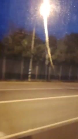 The video shows a blurred view of a road at dusk or night. A street light is visible on the left side of the frame, casting a bright beam. There is a road with white lane markings, and a dark fence runs parallel to the road, bordered by trees behind it. The sky is a deep blue. The perspective suggests the video was filmed from a moving vehicle.