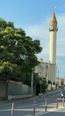 The video shows a mosque with a tall, cream-colored minaret topped with a golden, conical dome and a crescent moon. The mosque itself is a light beige building with multiple arched windows. Several golden domes are also visible on the roof of the mosque. The scene is set against a clear blue sky with a few wispy clouds. A large green tree is on the left side of the frame, and a metal fence runs along the property. A road is in the foreground with a dark blue car driving away from the camera. Several orange and white traffic cones are placed along the side of the road. The video appears to be taken from a moving vehicle.