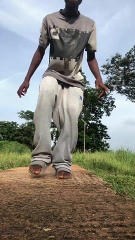 A young boy with dark skin and short black hair is captured in a video that appears to be recorded outdoors on a sunny day. He is wearing a grey t-shirt with a graphic print and light grey sweatpants. The boy is walking on a raised, dirt path surrounded by green grass and trees. He is looking at the camera and moving his arms as if dancing or performing a routine. His feet are bare, and he is wearing simple sandals. The background features a clear blue sky with some wispy white clouds. The video is shot from a low angle, emphasizing the boy's movement and the sky.
