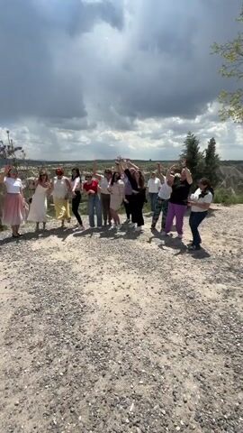 In a wide outdoor shot, a group of approximately 15-20 people, predominantly women, are gathered on a gravel and dirt patch, lively dancing or posing for a video. They are dressed in various casual to semi-casual outfits, including dresses, skirts, and pants in an array of colors like white, red, yellow, black, and purple. Most individuals have their arms raised, some swaying their bodies, indicating a festive and joyful atmosphere.

A man in a yellow shirt and khaki pants walks in front of the group, holding a mobile phone and appearing to film or interact with them, encouraging their actions. Another man in a white t-shirt and jeans is also seen among the group, participating in the posing. The background features a distinctive landscape with rolling hills and valleys, characteristic of a region like Göreme, Turkey, with sparse vegetation. The sky is partly cloudy with patches of blue, suggesting a daytime scene. Upbeat music is playing, enhancing the celebratory mood of the group.