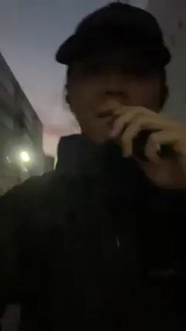 A person wearing a black baseball cap, a black jacket, and black gloves walks down an empty street bordered by buildings. The sky is a mix of grey and orange. The person appears to be vaping.