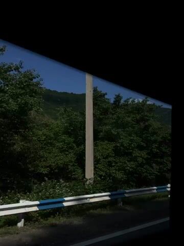 A view from inside a vehicle shows a green, tree-covered mountain range under a clear blue sky. The vehicle is moving, as indicated by the slight blur of the trees. A white and blue guardrail is visible along the side of the road.