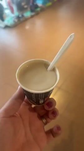 The video features a close-up shot of a hand holding a small paper cup filled with a light brown, creamy liquid, likely a dessert or beverage. A white plastic spoon is inserted into the cup, angled towards the viewer. The hand has bright pink nail polish and a silver ring on the ring finger. The cup has a black and white patterned exterior with some unreadable text.

The background appears to be an indoor setting with a tiled floor in a warm, light brown color. Further back, a display of colorful flip-flops on a rack is visible, suggesting a retail environment, possibly a shop or market. The lighting suggests it could be daytime, though no specific time can be determined. The overall mood is casual and perhaps leisurely, indicated by the act of holding and preparing to eat or drink something. The location is identified as Bertinoro, Italy.