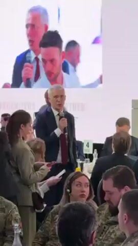 A video shows Jens Stoltenberg, Secretary General of NATO, speaking into a microphone during an event. He is standing near a table with a number marker, addressing an audience. The audience includes people in military uniforms and civilian attire. A woman in a green suit is looking at her phone. A camera is pointed at Stoltenberg. A screen shows a zoomed-in view of Stoltenberg and another man. Text on the screen behind Stoltenberg includes the words “ORGANIZERS & PARTNER.”
