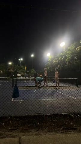 A group of people are playing pickleball on an outdoor court at night. The court is lit by overhead lights, and there are palm trees in the background. The people are wearing athletic clothing. One person is serving the ball, while others are positioned to return it. A fence surrounds the court.