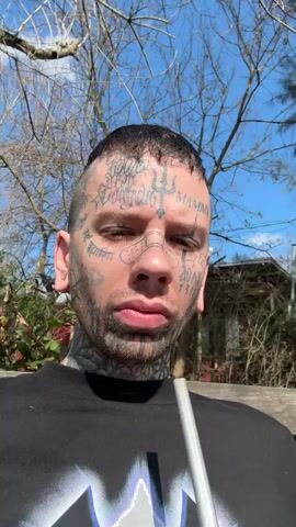 A person with facial tattoos and piercings sits on a wooden bench outdoors. They are wearing a black t-shirt with a graphic design and holding a metal straw in their mouth, which is inserted into a gourd filled with yerba mate. The background shows a grassy area with trees, a canoe, and a purple gas tank. The sun is shining, casting shadows.