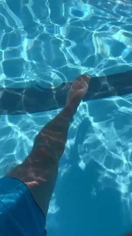 The media file shows an underwater view of a person's legs and feet in a swimming pool. The person is wearing blue swimming shorts. The water is clear and a dark line, possibly the edge of the pool or a lane marker, is visible running horizontally across the frame. Sunlight filters through the water, creating rippling patterns of light and shadow on the pool's surface and the person's legs. The overall impression is one of a bright, sunny day, suggesting a leisure activity like swimming. The location context of Vicenza, Italy, adds a potential geographical element to the scene. No text is visible in the media file.