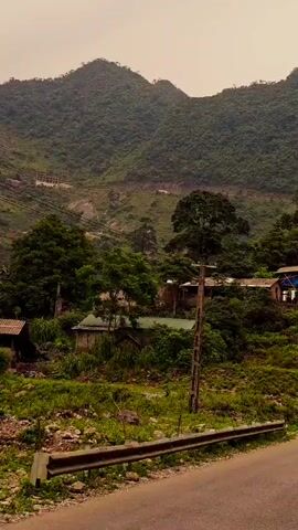 The camera is traveling along a paved road that curves through a lush, mountainous landscape. The mountains are covered in dense green forest, which extends down the slopes to the side of the road. There are several small buildings nestled among the trees. The sky is overcast, giving the scene a muted, somewhat hazy appearance. Utility poles run alongside the road, and a guardrail is visible in the foreground. There are also road signs visible along the road.