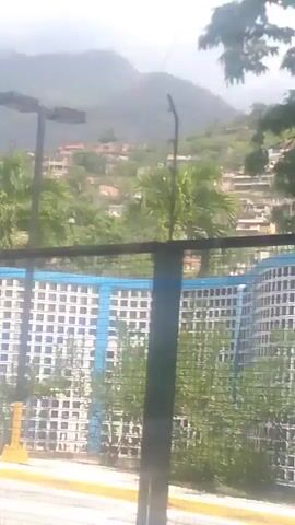 The video shows a panning view of an urban area with buildings on a hillside. In the foreground, there is a blue and white patterned fence and some vegetation. There are also shipping containers visible in the distance, some with the Hamburg Süd logo. The overall impression is of a port or industrial area located near a residential hillside.
