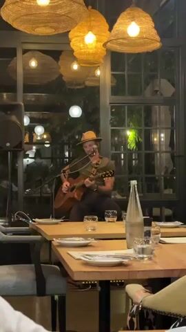 A video captures a male musician seated and playing an acoustic guitar in a restaurant setting. He wears a fedora-style hat, a green T-shirt, and dark pants. A microphone stands in front of him, suggesting he is singing, and a speaker is placed to his left. The restaurant has a modern aesthetic, with wooden tables set with plates, glasses, and water bottles.  Several wicker-style pendant lights hang from the ceiling, providing warm illumination.  The windows offer a glimpse of outdoor greenery.