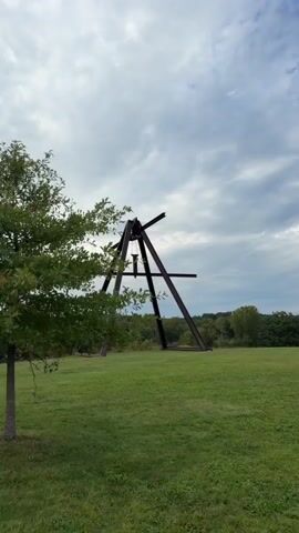 A wide shot of an outdoor sculpture is captured from a low angle. The sculpture is made of dark metal and has an A-frame structure with horizontal beams. It is situated on a grassy hill with trees in the background under a cloudy sky. The camera pans slightly to the right.