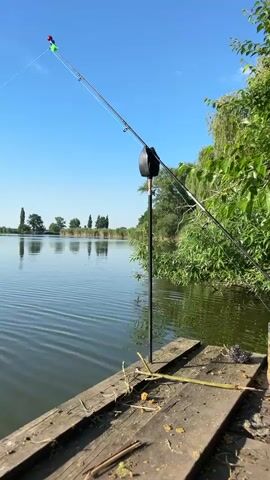 A fishing rod is propped up on a wooden dock near a lake. The fishing rod has a float on the end of the line. A can of beer is placed on the dock next to the fishing rod. The lake is calm and reflects the blue sky and trees on the opposite bank. The sun is shining and the sky is clear. The video is shot from a low angle, looking out over the water.