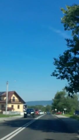 The video shows a road scene from a moving vehicle. The sky is clear blue. On the left side of the road, there is a yellow house with a brown roof and a balcony. There are trees and hills in the background. The road has double white lines in the center. Several cars are visible on the road, driving in the same direction as the camera. One black SUV is in the foreground on the left. Further down the road, there are red, white, and silver cars. On the right side of the road, there is a sidewalk and more trees. A red signpost is visible on the right.