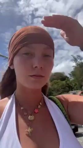 A young woman is filming herself by the coast on a sunny day. She wears a white top and a brown headscarf, and a necklace with sea-themed charms. Her expression is serious. She adjusts the headscarf while walking. The background includes palm trees, buildings, cars, other people, and the ocean.