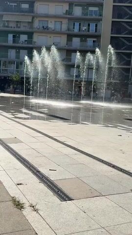 A public square features a water fountain with multiple jets spraying water upwards. The ground is paved with square tiles, and metal grates run parallel to each other across the paved area. In the background, a multi-story building with balconies and apartments is visible. The scene is illuminated by daylight.