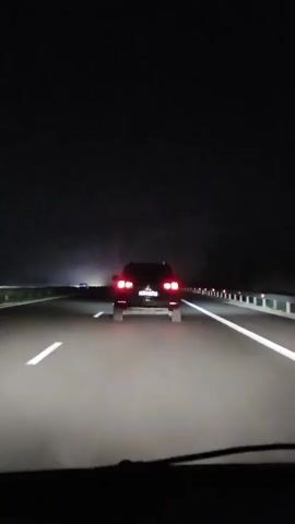A car is driving at night on a highway. The car in front is dark colored and its brake lights are illuminated. Headlights from oncoming traffic can be seen in the distance on the left side of the road. The road has white lines marking the lanes. The sky is dark, indicating it is nighttime.