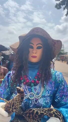 A person in a costume is dancing outdoors. The costume includes a mask with a gold face and dark eyes and lips, a brown floppy hat, a long-sleeved blue sequined top, a multi-colored beaded necklace, and a long white and blue skirt. The person is holding a stuffed animal that resembles a leopard or tiger. To the right of the person in costume, another person is dancing, wearing a dark suit, a black tie, a white shirt, and gloves. They also appear to be wearing a mask or heavy makeup that makes their face pale and their hair blond. The background shows a dirt road with some buildings and trees, and a few other people are visible in the distance. The sky is cloudy.