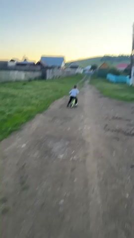 The video shows a person riding a bicycle down a dirt road in a village. The sun is setting, casting a warm glow on the sky and the surrounding houses. The houses are simple, wooden structures with colorful fences and roofs, indicative of a rural Russian setting like Aumyshevo. The road is lined with grass and trees, and a power pole with wires stands on the right side of the frame. The person on the bicycle is wearing a light-colored shirt and dark pants, with their back to the camera. They appear to be a child or young teenager. The overall atmosphere is peaceful and serene, capturing a quiet moment in village life at dusk. The video does not contain any visible text.