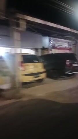 The video shows a nighttime view of a street scene. On the left, a dark-colored SUV is parked in front of an open garage or storefront. The vehicle has its rear facing the camera. Behind the SUV, a sign with blurry text is visible. A red object, possibly a cone or a bucket, is on the ground near the garage opening.

Further down the street, a small building with a pink and white facade is illuminated by bright lights. A scooter is parked in front of this building, and there are some red items on the ground near it. A white and red flag, possibly a national flag, is seen waving from a pole attached to one of the buildings.

The environment appears to be unpaved, with a dirt or gravel road. The overall lighting suggests it is nighttime, with artificial lights illuminating parts of the scene. The camera is in motion, causing some blurriness in the footage.