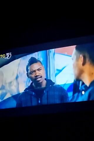 A man with a unique hairstyle and a plaid jacket is shown talking to another man. The scene then shifts to a different man, likely the same man from the previous scene, driving a car. He is wearing a blue button-down shirt. The video appears to be a clip from a movie or television show, with a channel logo in the corner. The environment is dimly lit, suggesting it is nighttime or indoors.