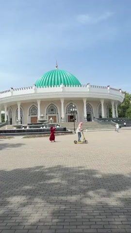 The video captures an outdoor scene featuring a grand, white circular building topped with a vibrant green dome and a golden finial. The building, possibly a museum or mausoleum, exhibits classical architectural elements with numerous columns, ornate arches, and intricate geometric patterns. In front of the building are several fountains and lampposts.

The ground is paved with light-colored bricks, forming an open plaza where several people are present. A person in a red garment is walking towards the left side of the frame, while another individual, wearing a light head covering, is riding a yellow kick scooter from right to left. Other figures are seen walking near the building's entrance in the background.

The weather appears sunny with a clear blue sky, casting distinct shadows on the plaza. Part of a person's arm and shoulder are visible in the top left and right foreground, suggesting a handheld camera perspective. The architecture is consistent with Central Asian styles, aligning with the given location of Uzbekistan. No visible text is present in the video.