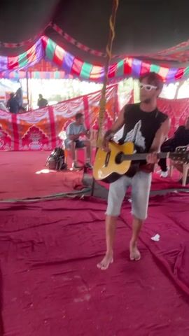 A man playing an acoustic guitar is seen in this short video.

The man is wearing sunglasses, a black muscle shirt with a white design, and jean shorts. He is barefoot and standing on a red rug. He appears to be singing while he plays.

There are other people seated and standing in the background, and they are watching the man play his guitar. They appear to be inside of a tent that has colorful decorations. A speaker is visible on the floor.