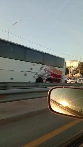 The video shows a view from inside a vehicle, likely a car, as it travels on a highway. The camera is positioned to capture the view out the side window and a reflection in the side mirror. On the highway, there are multiple vehicles, indicating traffic. Cars, trucks, and what appears to be a cement mixer are visible. The sky is clear with the sun low in the sky, suggesting it is either sunrise or sunset. The sun's reflection is prominent in the side mirror, casting a warm glow. Along the side of the highway, there are trees and some buildings or signs in the background. The overall impression is a snapshot of a commute or journey on a road with moderate traffic during a pleasant time of day.