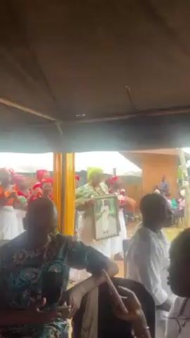 The video shows a group of people gathered under a canopy, with some women in the foreground dancing and singing. Most of the women in the group are wearing red tops and white bottoms with red headwraps. One woman in the front is carrying a framed photo, and another is playing an instrument. The people in the foreground are seated and watching the group of women. The environment appears to be outdoors, possibly a courtyard or garden area. The lighting is subdued under the canopy, but there is daylight visible outside.