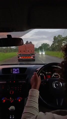 A person is driving a Mazda car on a wet road, following a truck loaded with bricks. The sky is overcast. Another car is approaching in the opposite lane. The interior of the Mazda is visible, including the dashboard and steering wheel. The driver's right hand is visible on the steering wheel.