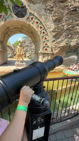 A girl is aiming a black cannon at a boat full of people on the water. The boat is moving towards the girl. There is a golden statue of a person under an archway. The water is green. The background is rocky.