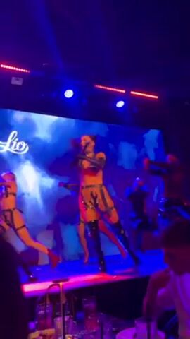 Here is a description of the video:

The video shows a live dance performance on a stage, presumably in a nightclub.  Three dancers are visible on the stage, wearing similar revealing costumes of tops, bottoms, and thigh-high boots.  The costumes have a leather-esque look with dark trim and accents.  The dancers are moving in a choreographed manner.  

The stage is elevated and features blue lighting and a large video screen behind the performers displaying the club's name "Lio."  The audience can be seen at tables in the foreground, watching the performance.  Several drinks are visible on the tables.  The camera angle is from the perspective of an audience member.
