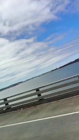 A video taken from inside a vehicle traveling over a bridge. The camera is pointed at an angle, capturing the sky, clouds, trees, and the body of water below. A guardrail is visible along the side of the road. The bridge structure itself can be seen in the distance. The overall impression is of a scenic drive on a partly cloudy day.