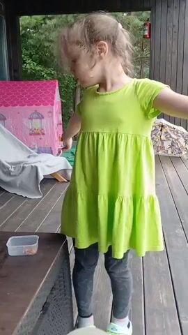 A young girl with blonde braided hair, wearing a bright green tiered dress and grey leggings, stands on a wooden deck. She is facing to the left, looking down at a small clear plastic container on a dark wicker table. She appears to be clapping her hands together. In the background, there is a pink play tent with a yellow slide, trees, and a wooden structure.