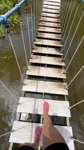 A first-person perspective video shows a person's legs and feet as they walk across a narrow, wobbly wooden rope bridge over murky water. The bridge is constructed of weathered wooden planks suspended by ropes on either side. The person is wearing black shorts and bright pink sandals. The water below the bridge appears dark and still, with some green aquatic plants visible along the banks. The scene suggests an outdoor, possibly park-like or natural setting, with lush greenery and trees visible in the background. The video captures the sensation of movement and the slightly precarious nature of crossing the bridge.