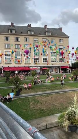 A wide shot captures a bustling outdoor plaza. In the background, a multi-story building is adorned with numerous colorful hot air balloon decorations strung across its facade. The plaza itself features a manicured green lawn with several people relaxing in lounge chairs. Beyond the lawn, a tiered set of stone steps leads up to a row of trees and outdoor seating areas, likely for cafes or restaurants. In the foreground, a water feature sprays mist into the air, with a young child standing near it. The overall scene suggests a lively, public space on a cloudy day.