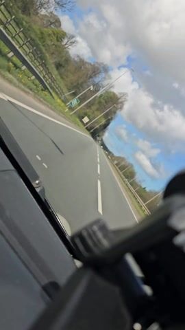 A dashboard-level view from a car driving on a road. The sky is blue with white clouds. Green trees line the side of the road. There is a sign to the left of the road. There is a double white line in the middle of the road. Other cars are on the road.