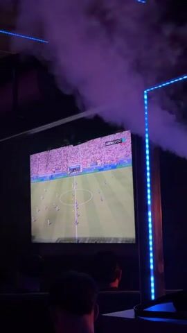 The video shows a large screen displaying a soccer match. The screen is set up in a dark room with purple and blue lighting. Smoke or fog is visible in the air, obscuring the top of the screen. Two people are seated in the foreground, with their backs to the camera, watching the game. One person's head is visible from the shoulders up. The soccer field is green with white markings, and there are players in blue and white uniforms on the field. The stands are filled with spectators wearing pink and white. A logo for "ARENASPORT" is visible in the upper right corner of the screen. The lighting in the room is atmospheric, with blue LED lights lining a corner or structure to the right of the screen.