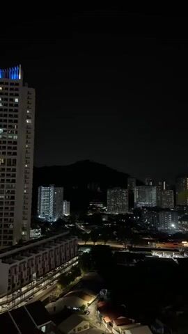 A vertical night photograph captures an expansive urban landscape, likely George Town, Malaysia, viewed from an elevated perspective. The sky is dark and clear, typical of nighttime. In the immediate foreground on the left, a prominent, tall white high-rise building with numerous lit windows stands, topped with a distinctive blue light feature. Below it, a section of lower-rise residential buildings with red-tiled roofs and visible balconies extends across the bottom left.

The midground reveals a dense city scene with various buildings, predominantly residential high-rises and apartment blocks, many of which are illuminated, creating patterns of light across the urban fabric. Streaks of light on the roads below indicate vehicle movement. Trees and dark foliage are scattered throughout the cityscape. In the background, a dark, silhouetted mountain or hill range rises against the night sky, providing a natural boundary to the urban sprawl. No people or visible text are present in the image. The scene conveys the quiet and expansive glow of a city at night.