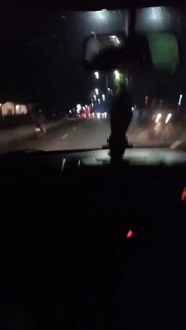 The video is shot from inside a car at night, looking out through the windshield. The road ahead is lit by streetlights and the headlights of other vehicles. To the left of the road, there is a sidewalk and some buildings or structures. A person on a motorcycle is visible on the left side of the road, moving away from the camera. In the center of the frame, hanging from the rearview mirror, is a small decorative object. The dashboard of the car is visible in the foreground, with some lights illuminated. The overall scene depicts a nighttime drive through a populated area.