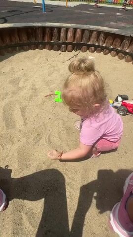A blonde-haired toddler with her hair in a bun is playing in a sandbox. She is wearing a pink t-shirt and pink shorts and pink shoes. She is crouching on her knees and drawing in the sand with her finger. There are toys in the sandbox, including a red and white toy truck. The sandbox is surrounded by a wooden border and is part of a playground. The ground in the playground is covered with red and black rubber mulch. There are colorful playground equipment in the background. The sun is shining, casting shadows on the sand.