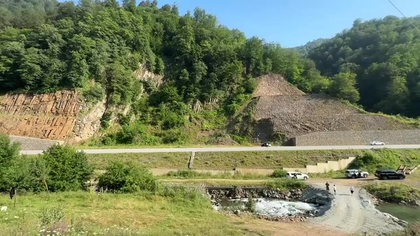 A wide shot captures a mountainous landscape under a clear sky. A paved road cuts horizontally across the scene, with a steep, wooded mountainside rising behind it. The mountainside features exposed rock formations on the left and a large, cleared area of loose rock and gravel in the center-right, appearing to be a slope reinforcement or construction zone. A stream with turbulent water flows in the foreground, crossed by a rudimentary bridge. Several vehicles are present: two white SUVs, a black SUV, a black truck, and what appears to be construction equipment with a long arm on the right side of the road. A few individuals are visible near the stream and the road's edge. The vegetation is lush green, indicating a natural, undeveloped environment.