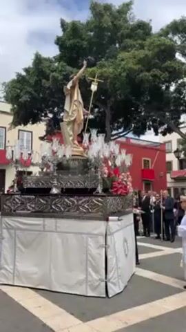 The video captures an outdoor religious procession in a paved square. A large, ornate silver float is prominently featured, adorned with numerous silver candlesticks, red and white flowers, and a statue of Jesus Christ holding a cross, arm raised. The float is covered with a white fabric skirt. Surrounding the float are various people, including men in suits, a woman in a white cleric's robe, and other onlookers. The background includes buildings with red and white facades and large trees under a partly cloudy sky. Loud music, likely from a brass band, is playing.
