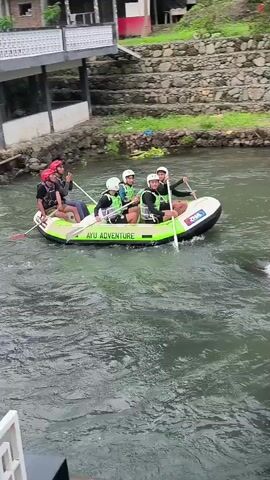 Five people are in a white and green inflatable raft on a river. The raft has "AYU ADVENTURE" written on its side. The people are wearing life vests and helmets, and four of them are holding paddles. The raft is moving through white water. The river is surrounded by a stone wall and some greenery.