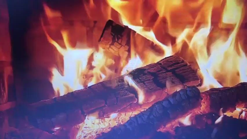 A close-up shot shows a fireplace with burning logs. The flames are orange and yellow, rising and dancing around the logs. The logs are dark and charred, with visible cracks and textures. The background is brick, suggesting the interior of a fireplace. The scene is static.