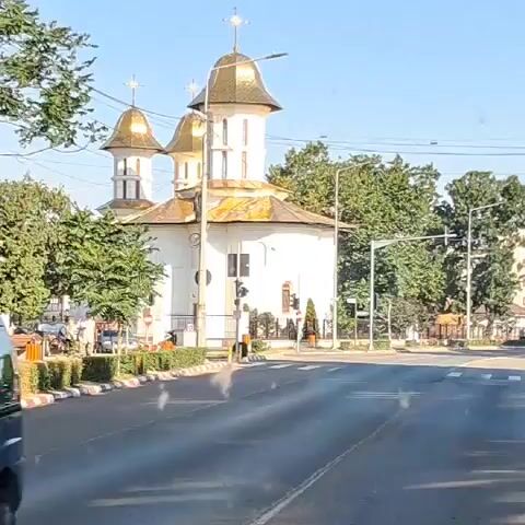 A white church with gold domes is visible on the left side of the road. A traffic light is also present. Cars are driving on the road, and there are trees and buildings in the background. The video shows a slight upward angle towards the sky. In a later frame, a geyser-like plume of water erupts in the middle of the intersection, with cars stopped around it. Another frame shows the intersection clearing up, with traffic resuming. The last frame shows cars driving down a straight road lined with trees and a sidewalk.