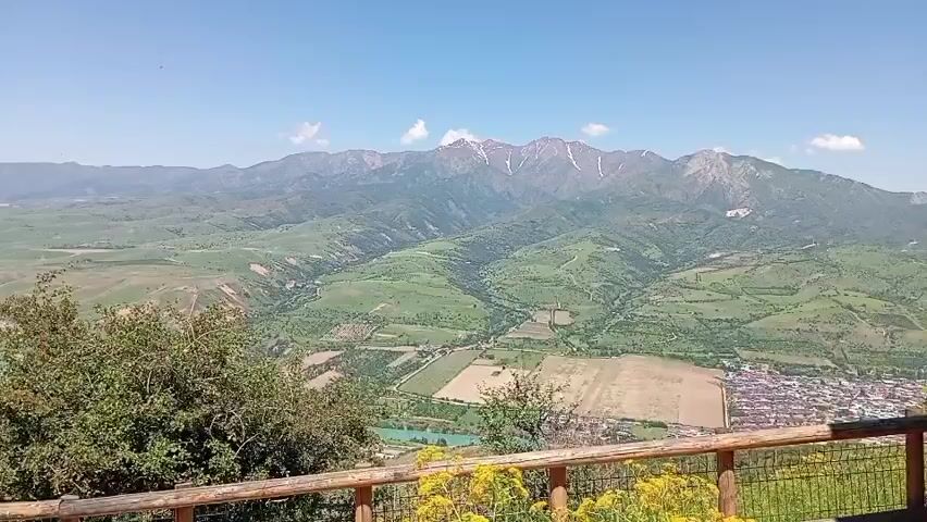 The video shows a wide shot of a mountainous landscape with a river running through a valley. The valley is mostly green with some patches of farmland and what appears to be a small town. The mountains in the background have snow-capped peaks. In the foreground, there is a wooden fence and some foliage. As the video progresses, the camera pans slightly to the left, revealing a person standing on a rocky outcropping. The video also shows some tree branches with leaves hanging in the top of the frame. The sky is blue and mostly clear. The overall tone of the video is peaceful and serene.